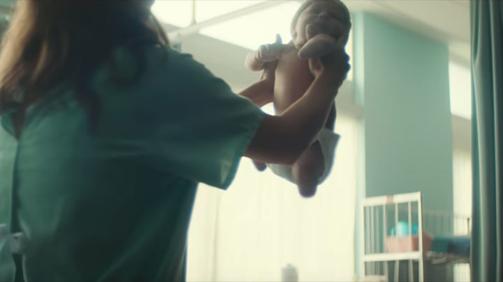 Image of a woman in a hospital robe holding up a baby with 3 missing limbs. The baby is wearing a diaper and the woman matches the Hospital Green paint and curtains on the wall.