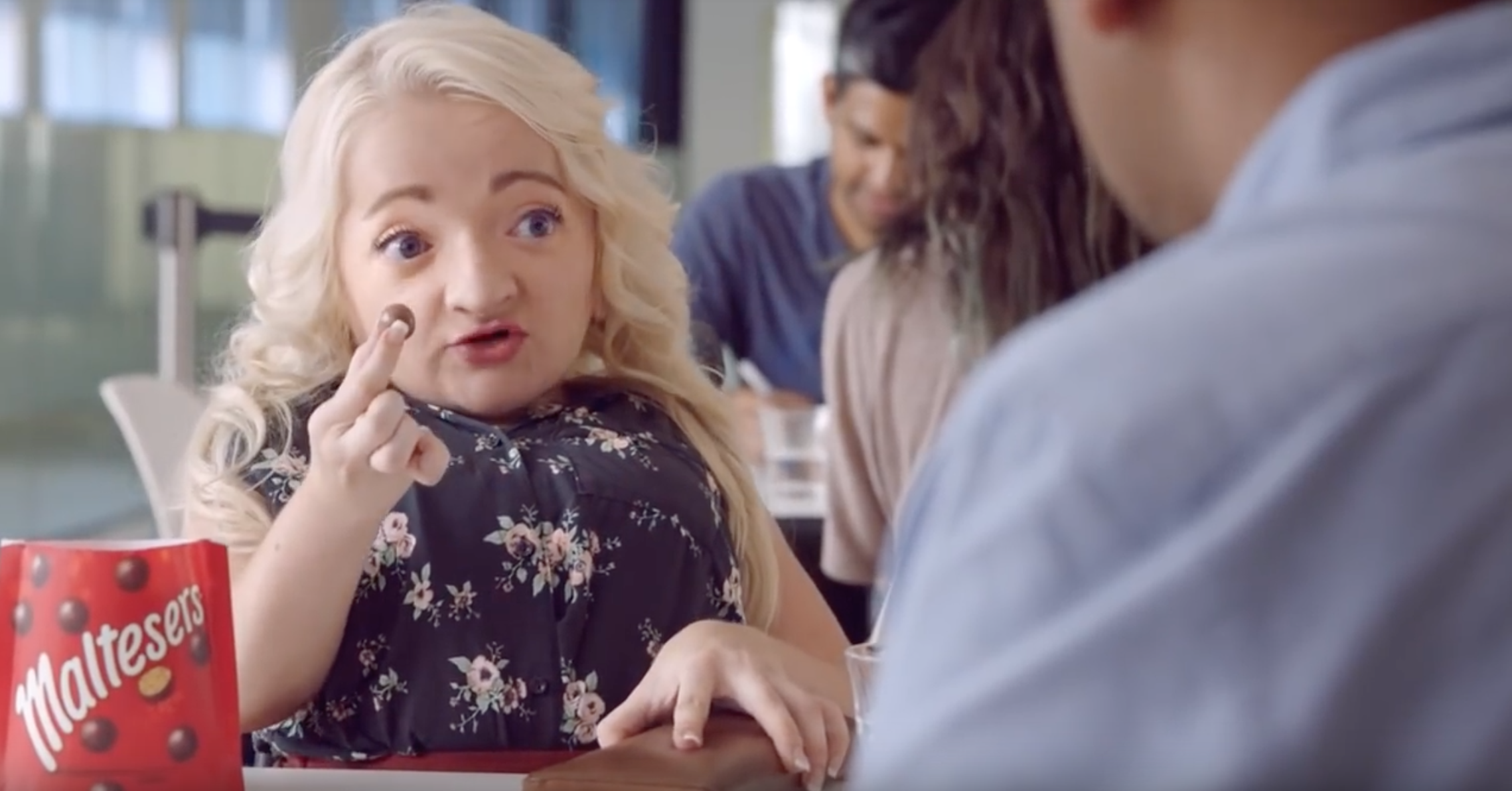 Image of a white woman with platinum blonde hair holding up a piece of Maltesers, with the Maltesers packing in the foreground, while she talks to the person, who is off screen, across from her. They are seated at a lunch table.