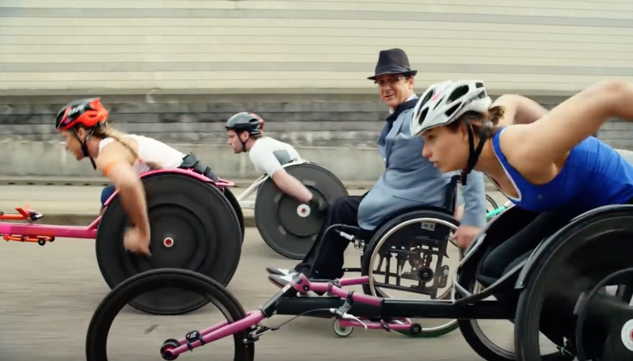 Image of two white woman and one white man going full speed in racing wheelchairs while a white man in a fedora and and a manual wheelchair and a suit looks at the camera while moving at a casual pace.