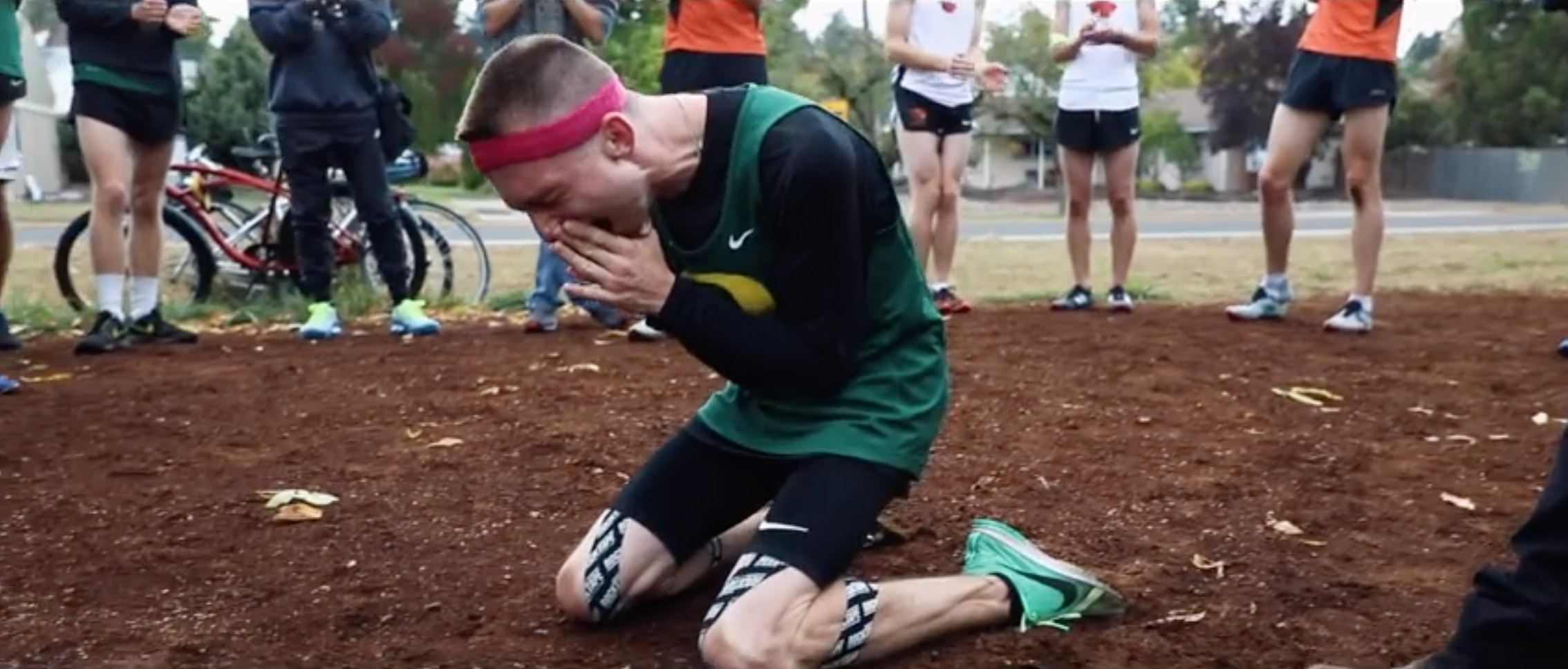 Image of a white man, college age, wearing a green Oregon State jersey and running gear, kneels on the dirt ground, hands to his face while he sobs.