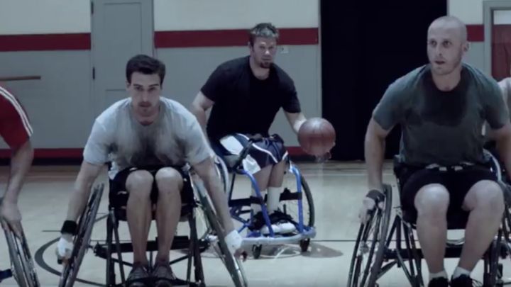 Image of a group of 5 men, all white, all sweaty wheeling to the other end of the court in wheelchair basketball. The man in the middle is dribbling the ball.