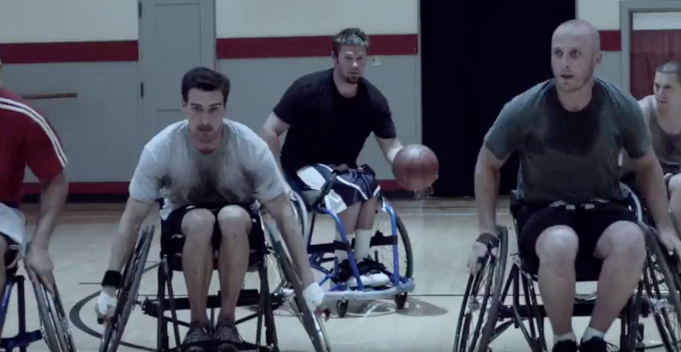 Image of a group of 5 men, all white, all sweaty wheeling to the other end of the court in wheelchair basketball. The man in the middle is dribbling the ball.