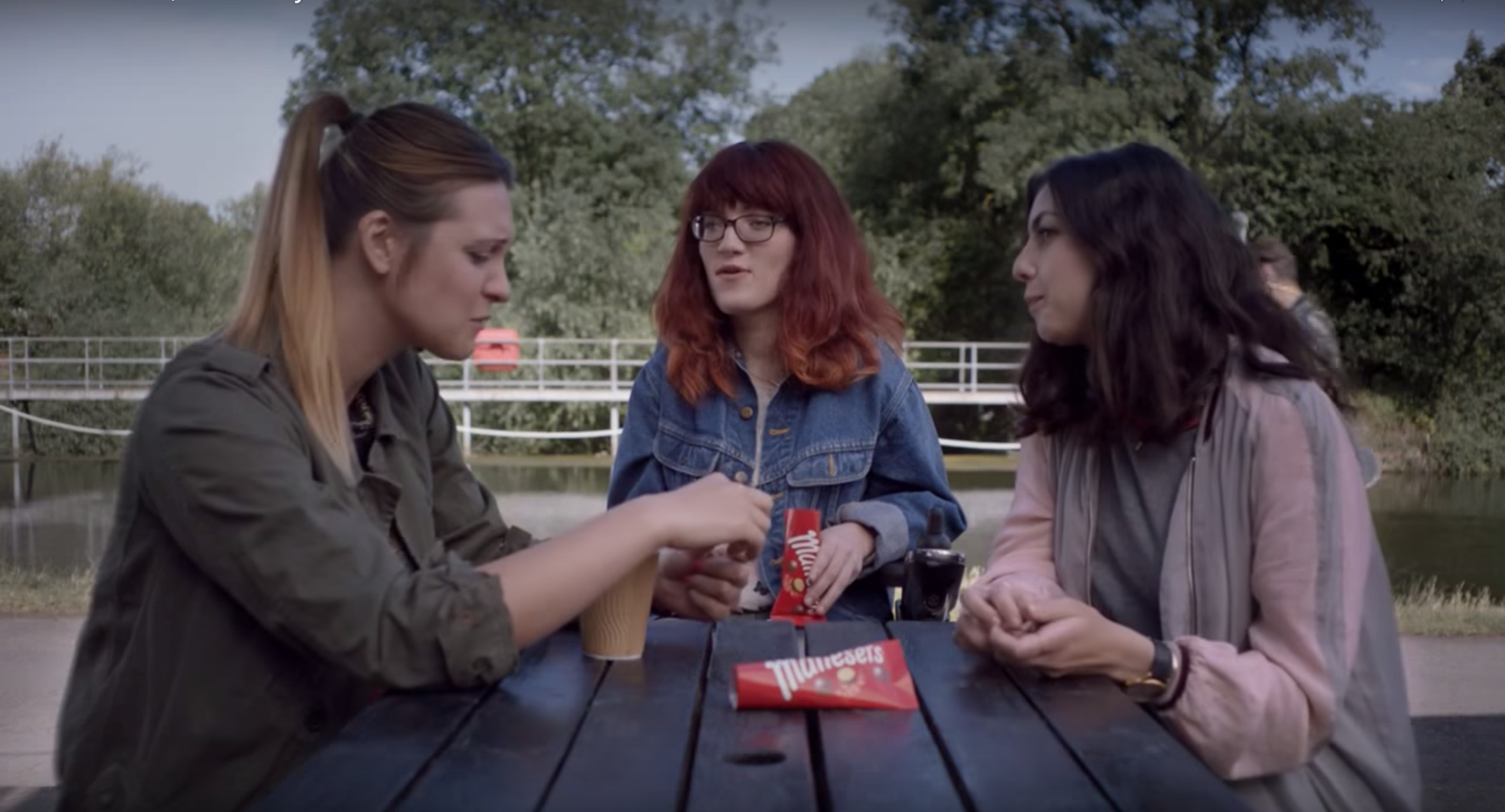 Image of Red Headed woman with red hair and glasses in a wheelchair talking to two friends at a picnic bench while eating Maltesers
