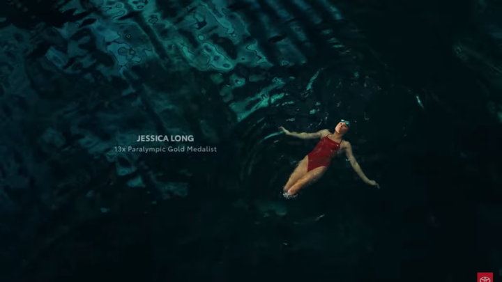 Image from the beginning of the ad. Jessica Long, a double amputee wears a red swimsuit while swimming in dark water. White text on the screen tells us she's a 13x paralympic gold medalist
