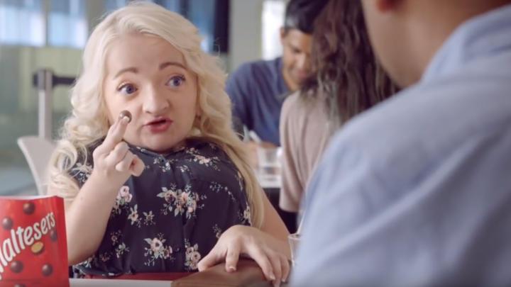 Image of a white woman with platinum blonde hair holding up a piece of Maltesers, with the Maltesers packing in the foreground, while she talks to the person, who is off screen, across from her. They are seated at a lunch table.