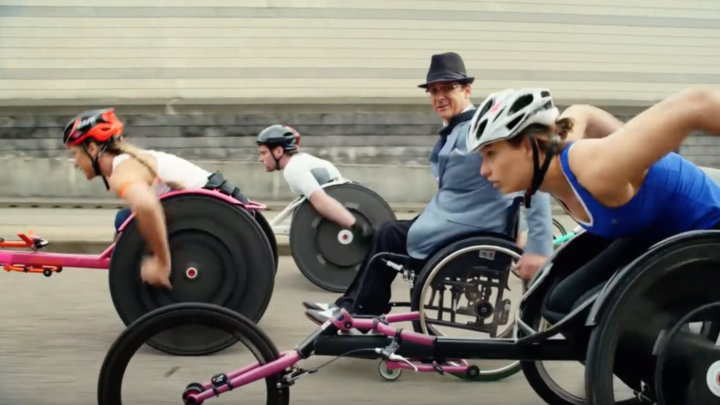 Image of two white woman and one white man going full speed in racing wheelchairs while a white man in a fedora and and a manual wheelchair and a suit looks at the camera while moving at a casual pace.