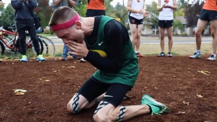 Image of a white man, college age, wearing a green Oregon State jersey and running gear, kneels on the dirt ground, hands to his face while he sobs.