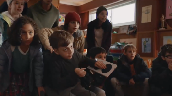 Image of a group of a dozen kids sitting in a den, all staring intently at a screen, while the boy in the middle uses the Xbox Adaptive Controller to play.