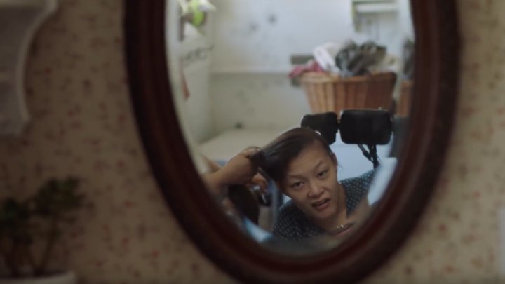 Image of an asian woman in a wheelchair looking into a camera while another woman, who is almost entirely out of frame pulls her hair into a pony tail.