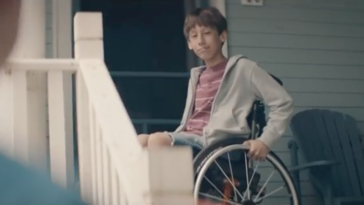 Image of a young white boy, perhaps ten years of age, sitting in a wheelchair on a porch that's up a few steps, while another boy his age looks on from the driveway.