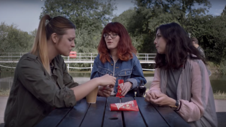 Image of Red Headed woman with red hair and glasses in a wheelchair talking to two friends at a picnic bench while eating Maltesers