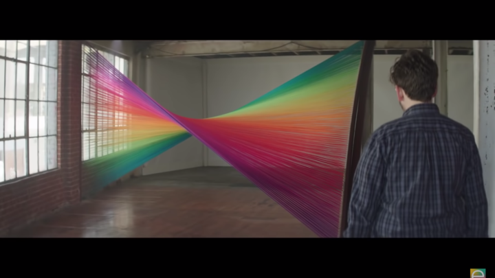 Image of a man standing in front of a large string rainbow colored installation inside a brightly lit room